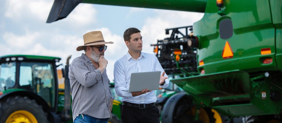 Professional farmer with a modern combine at field in sunlight at work. Confident, bright summer colors. Agriculture, exhibition, machinery, plant production. Senior man near his tractor with investor.